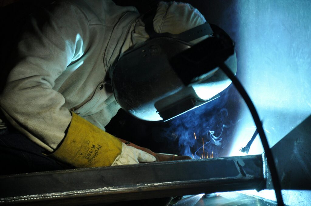 A welder working indoors, creating sparks, wearing safety gear in an industrial setting.